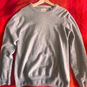 Grå stickad tröja från GANT i superfin lammull  - Säljer en stilren grå stickad tröja från GANT i superfin lammull. Klassisk rund halsringning, ribbade muddar och diskret broderad logga på bröstet. Perfekt för lager-på-lager och chill höst- eller vinterstil.