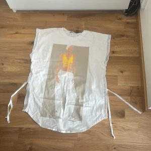 Vit t-shirt med tryck från Zara - Vit t-shirt från Zara med stort grafiskt tryck framtill av en person i gult och vitt. T-shirten har ärmlös design, mesh-detaljer längs sidorna och snörning i midjan. Oversized och loose fit, tillverkad i mjuk bomull.