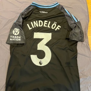 Aston Villa Lindelöf #3  - Snygg svart Aston Villa fotbollströja från Adidas med Lindelöf och nummer 3 på ryggen. Tröjan har ljusblå detaljer på axlar och krage, klubbmärke på bröstet och sponsorer på ärm och framsida. Tillverkad i lätt och ventilerande material.