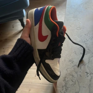 Nike Air Jordan 1 Low Multicolor - Säljer ett par Nike Air Jordan 1 Low med färgglad design. Skorna har vit bas, svarta detaljer, röd swoosh, blå och gröna paneler samt orange foder. Klassisk rund tå, svart snörning och svart sula. Perfekta för dig som vill sticka ut med dina sneakers.