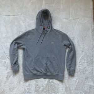 Grå hoodie från BOSS med snörning - Grå hoodie från BOSS med klassisk känguruficka och snörning i huvan. Tröjan har långärmade muddar och en liten BOSS-logga på bröstet. Tillverkad i mjuk bomull, perfekt för chill dagar. Snygg och enkel design som funkar till allt.