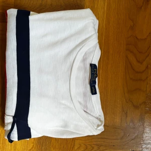 Randig långärmad tröja Polo Ralph Lauren - Vit långärmad tröja från Polo Ralph Lauren med röda och marinblå ränder. Tröjan har en liten bröstficka och broderad logga nertill. Tillverkad i mjuk bomull, perfekt för en chill och stilren look.