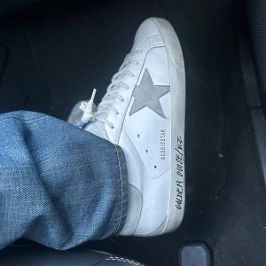 Golden Goose Superstar  - KVITTO OCH ALLT OG FINNS!!! Golden Goose Superstar sneakers i vitt skinn med grå mockadetaljer och ikonisk stjärna på sidan. Snygg text på sulan och blå etikett på plösen. Klassisk låg modell med snörning och platt sula, perfekt för en avslappnad och trendig look. 