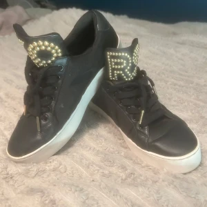 Svarta Michael Kors sneakers med pärldetaljer - Svarta sneakers från Michael Kors i skinn med vit platt sula. Tungan har stora guldiga pärlor som bildar texten 'KORS' och skorna har svarta snören med gulddetaljer. Normal i strl