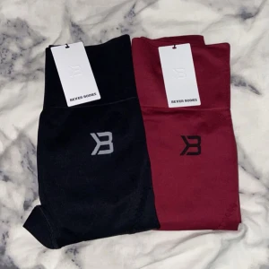 Better Bodies leggings i svart & vinröd - Två par snygga leggings från Better Bodies, ett par i svart med grå logga och ett par i vinrött med svart logga. Båda har hög midja och är tillverkade i stretchigt syntetmaterial, perfekta för träning. Detaljerad struktur på benen för extra stil.