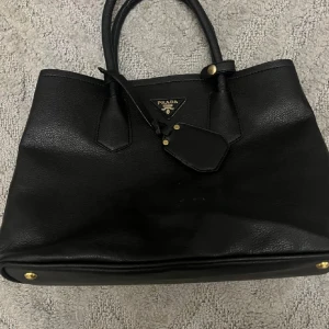 Svart handväska  - Snygg svart handväska med guldfärgade metalldetaljer och klassisk trekantig logotyp framtill. Väskan har dubbla handtag och en stilren, tidlös design som passar till många olika looks.