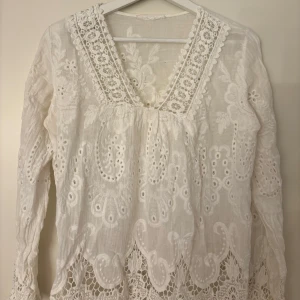 Vit broderad blus med spetsdetaljer - Superfin vit blus med broderade blommor och spetsdetaljer längs v-ringningen och nederkanten. Perfekt till sommaren eller våren! Vet ej vart den är ifrån men storleken är i passform till xs-s