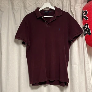 Vinröd pikétröja Polo Ralph Lauren L - Vinröd pikétröja från Polo Ralph Lauren i modellen Custom Slim Fit. Klassisk krage, två knappar och broderad logga på bröstet. Tillverkad i mjuk bomull och har korta ärmar. Perfekt för en clean och stilren look!