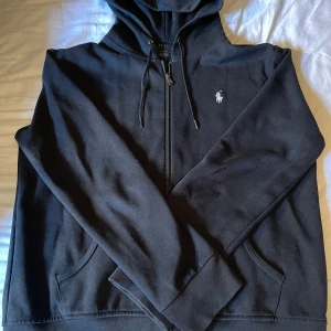 Svart hoodie från Polo Ralph Lauren - Svart hoodie från Polo Ralph Lauren med dragkedja framtill, snörning i huvan och klassisk logga broderad på bröstet. Tillverkad i mjuk bomull och har fickor på sidorna. Perfekt för chill dagar eller när du vill ha en clean look.   Hoodien är bara testad 1 gång och är i väldigt fin skick.