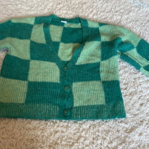 Grön rutig kofta från Lindex - Säljer en stickad kofta från Lindex i gröna nyanser med rutigt mönster. Koftan har v-ringning, tre knappar framtill och långa ärmar. Perfekt för dig som gillar färg och vill ha en mysig och trendig look.