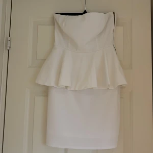 Vit axelbandslös peplumklänning - Snygg vit axelbandslös kortklänning med peplumvolang i midjan. Stilren och elegant design som ger en feminin siluett. Perfekt för dig som vill sticka ut med en klassisk look.