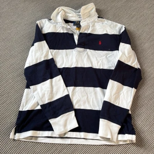 Ralph Lauren Polo (junior)  - Randig pikétröja - Säljer en klassisk långärmad pikétröja från Polo Ralph Lauren i marinblått och vitt. Tröjan har breda ränder, krage och knäppning vid halsen samt den ikoniska röda loggan broderad på bröstet. Tillverkad i mjuk bomull, perfekt för en chill och sportig look.