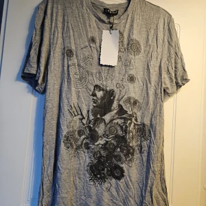 Grå t-shirt med konstnärligt tryck So Much - Unik grå t-shirt från So Much i storlek L med ett stort konstnärligt tryck framtill som blandar ansikte, blommor och grafiska detaljer. Materialet är mjukt och består av 50% bomull och 50% viskos. Rund hals och korta ärmar ger en avslappnad vibe.