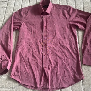 Rosa skjorta från Benezzo slim fit - Snygg rosa skjorta från Benezzo i slim fit-modell. Skjortan har klassisk krage, långa ärmar med manschett och knäppning framtill. Liten broderad logga på bröstet och matchande rosa knappar. Perfekt för dig som gillar stilrena plagg med färg.