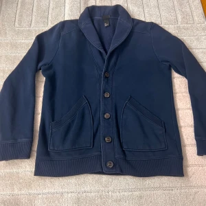 Mörkblå kofta med knappar och sjalkrage - Mysig mörkblå kofta från Uniqlo med sjalkrage och stora knappar framtill. Koftan har två rejäla fickor och är fodrad med mjuk fleece på insidan. Perfekt för kyliga dagar och enkel att matcha med det mesta.