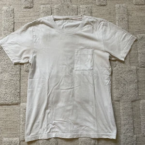 Vit t-shirt från Nudie Jeans - Vit basic t-shirt från Nudie Jeans med rund halsringning och en bröstficka. Tillverkad i mjuk bomull och har en klassisk passform. Perfekt för en clean och enkel stil.