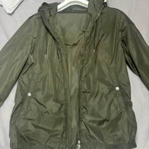 Olivgrön vindjacka från Moncler - Snygg olivgrön vindjacka från Moncler med huva och dragsko, klassisk logga på ärmen och röd-vit-blå rand på huvkanten. Jackan har dragkedja framtill, två fickor med tryckknapp och är tillverkad i glansig polyester. Perfekt för dig som vill ha en stilren och sportig look.