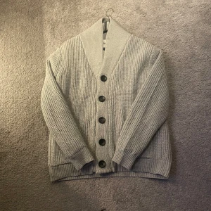 Grå stickad kofta med knappar - Säljer en grå stickad kofta med bred krage och stora svarta knappar framtill. Koftan har långärmad design och ribbade muddar vid ärmslut och nederkant. Perfekt för lager-på-lager och chill dagar.