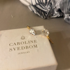 Guldarmband Caroline Svedbom kristall - Snyggt armband från Caroline Svedbom i guldtonad metall med tvinnad design och två stora, gnistrande klara kristaller i varje ände. Perfekt statement-piece för dig som gillar bling och exklusiv känsla.