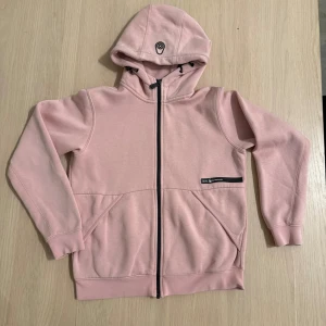 Rosa hoodie från Sail Racing med dragkedja - Ljusrosa hoodie från Sail Racing med svart dragkedja och logga på bröstet. Tröjan har huva med snörning, två fickor framtill . Perfekt för en chill och sportig stil. Storlek 170 men passar 160, 170, XXS