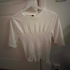 Vit smockad topp med volangkant - Vit topp med smockad midja och volangkant nertill. Toppen har rund halsringning och halvlånga ärmar och är tillverkad i mjuk bomull. Den är i storlek S och endast använd en gång så i väldigt bra skick.