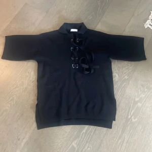 Svart tröja med snörning från Stefanel - Svart tröja från Stefanel med korta ärmar och cool snörningsdetalj framtill. Tröjan har en hög krage och är tillverkad i ett mjukt stickat material. Perfekt för dig som gillar en stilren look med en twist. Made in Italy