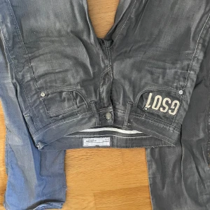 Grå jeans från G-Star RAW med tryck - Säljer ett par grå jeans från G-Star RAW med coola vita tryckdetaljer på bakfickorna och unika sömmar. Jeansen har klassisk femficksdesign, dragkedja och knapp framtill samt tydliga kontrastsömmar. Perfekta för dig som gillar streetwear och vill sticka ut.