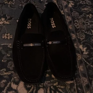 Svarta loafers från BOSS med metallspänne - Snygga svarta loafers från BOSS med stilren design och metallspänne framtill. Skorna har rund tå och dekorativa sömmar längs kanterna. Perfekta för dig som gillar klassisk men modern stil. Materialet är mocka för en lyxig känsla.