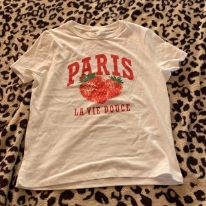 Vit t-shirt med jordgubbar från H&M - Vit t-shirt från H&M med röd text 'PARIS' och 'LA VIE DOUCE' samt tryck av två jordgubbar på bröstet. Klassisk rund hals och korta ärmar. Perfekt för dig som gillar fräscha och söta prints.