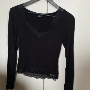 Svart långärmad topp med spetsdetaljer - Svart långärmad topp från Gina Tricot med v-ringning och fina spetsdetaljer längs halsringningen och nederkanten. Toppens passform är figurnära och materialet är mjukt och stretchigt, perfekt för en stilren look. Knappt använd.
