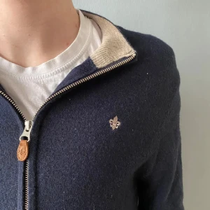 Marinblå kofta i merinoull från Morris - Snygg marinblå kofta från Morris i 100% merinoull. Dragkedja framtill med läderdetalj, beige insida på kragen och broderad logga på bröstet. Krympt lite i tvätten så sitter mer som M eller S.