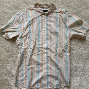 Randig kortärmad skjorta från Riley - Kortärmad skjorta från Riley i slim fit med fräscha vertikala ränder i ljusblått, beige och vitt. Skjortan har klassisk krage och knäppning framtill, perfekt för en chill sommardag. Tillverkad i mjuk bomull som känns skön mot huden.