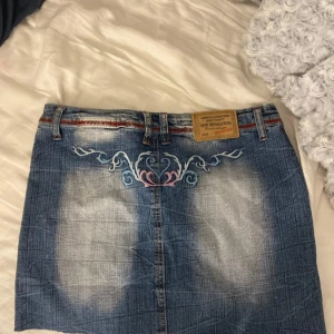 Jeanskjol med broderi från New Sensation - Snygg kort jeanskjol från New Sensation med slitna detaljer, röd dekorstickning och broderier i blått, rosa och vitt. Kjolen har bälteshällor, knapp och dragkedja framtill samt ett litet hjärta broderat på ena sidan. Perfekt för dig som gillar unik denimstil.
