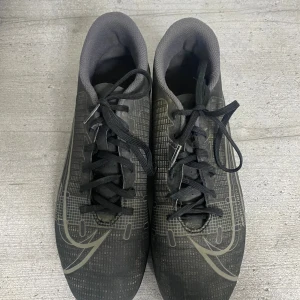 Nike Mercurial svarta fotbollsskor - Svarta Nike Mercurial fotbollsskor med silverdetaljer och snörning. Skorna har låg profil, platt sula med dobbar och är tillverkade i syntetmaterial. Snyggt grafiskt mönster på ovandelen och klassisk Nike-logga på sidan. Perfekta för dig som vill sticka ut på planen.