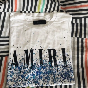 Vit t-shirt från Amiri med blått stänk - Snygg vit t-shirt från Amiri med stort svart tryck framtill och coolt blått färgstänk-mönster längst ner. Klassisk passform och rund halsringning. Perfekt för dig som vill sticka ut med en unik design.