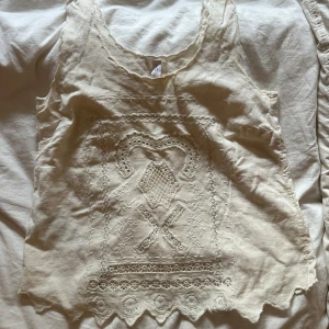 Beige linnetopp med broderi och spets - Supersöt beige linnetopp med broderade detaljer och spets längs nederkanten. Toppen är ärmlös och har en rundad halsringning. Perfekt för en romantisk och bohemisk look.❤️🥰