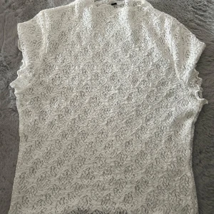 Vit spetsblus med kort ärm - Säljer en vit blus i spets med vackert mönster och korta ärmar. Toppen har en hög hals och är tillverkad i ett tunt, genombrutet material som ger en romantisk känsla. Perfekt att styla med jeans eller kjol för en trendig look.
