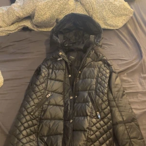 Svart quiltad dunjacka Rockandblue - Svart dunjacka från Rockandblue med snyggt quiltat mönster, huva och dragkedjor på fickorna. Jackan har en normal passform och är perfekt för kalla dagar. Märke på ärmen och detaljer i silver. Stängs med både dragkedja och knappar. Den passar både S och M 