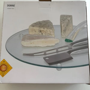 Cheese Tray - Elegant ostbricka från Dorre med rund glasskiva och tillhörande ostknivar. Perfekt för servering av olika ostar vid fest eller middag. Produkten är i nyskick och levereras i originalförpackning med skyddande bubbelplast. Inga synliga defekter eller slitage.