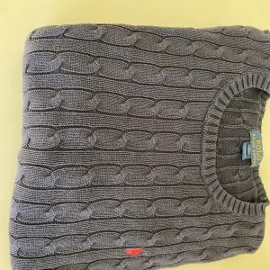 Mörkblå kabelstickad tröja Polo Ralph Lauren - Kabelstickad mörkblå tröja från Polo Ralph Lauren med rund halsringning och klassisk röd broderad logga på bröstet. Tröjan är långärmad och har ribbade muddar vid ärmslut och nederkant. Perfekt för dig som gillar stilren och tidlös design. Pris kan såklart diskuteras! 