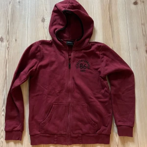 Vinröd hoodie från Peak Performance - strl.S - Vinröd hoodie från Peak Performance med dragkedja framtill, huva och två fickor. Tryck med '86' och logga på bröstet. Skön och mjuk i bomullsmix, perfekt för kyliga dagar. Klassisk passform och ribbade muddar vid ärmslut och nederkant.