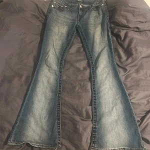 True Religion bootcut jeans med röd söm - Säljer ett par blå bootcut jeans från True Religion med tydliga röda kontrastsömmar och klassiska bakfickor med broderi. Jeansen är low raist och är tillverkade i denim med slitningar och ljusare partier för en cool vintage-look.
