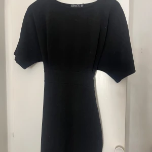 Svart kortklänning från Boohoo - Svart kortklänning från Boohoo med breda korta ärmar och rund hals. Klänningen har ett snyggt knytband i midjan som ger en fin siluett. Enkel och stilren design, perfekt för dig som gillar minimalistiskt mode.