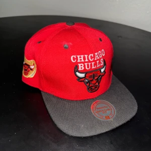 Chicago Bulls keps från Mitchell & Ness - Snygg röd Chicago Bulls snapback-keps från Mitchell & Ness med svart skärm och broderad logga framtill. Kepsen har justerbar passform, NBA-märke på sidan och broderad Mitchell & Ness-text baktill. Perfekt för dig som gillar basket och streetwear.