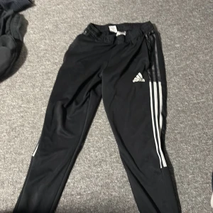 Svarta Adidas träningsbyxor med ränder - Svarta träningsbyxor från Adidas med klassiska vita ränder längs sidorna och Adidas-logga på låret. Ni står för frakt!