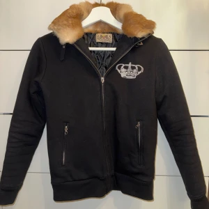 Vintage Svart jacka Juicy Couture med päls - Svart jacka från Juicy Couture med broderad krona på bröstet och texten 'Crimes Of Couture' i rosa på ryggen. Jackan har dragkedja, två fickor med dragkedja och en avtagbar krage i brun päls. Fodret är quiltat för extra värme. Vänster ficka går ej att öppna pga att dragkedjan är fast och det saknas ett J på den stora dragkedjan. Annars knappt använd. Medium egentligen men liten i storlek.