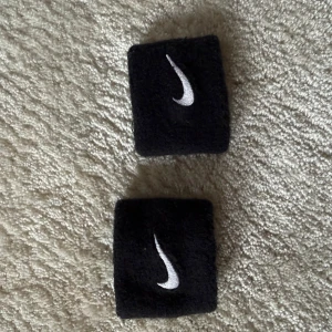 Svarta Nike svettband - Ett par svarta svettband från Nike med klassisk vit swoosh-logga broderad på framsidan. Tillverkade i mjukt och absorberande frottématerial, perfekta för sport och träning. Sitter tajt runt handleden och ger en sportig look.