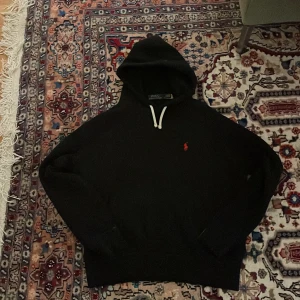 Svart hoodie från Polo Ralph Lauren - Svart hoodie från Polo Ralph Lauren med klassisk röd broderad logga på bröstet. Tröjan har huva med vit snörning och är tillverkad i mjuk bomull. Perfekt för en chill och stilren look.