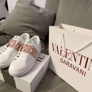Valentino Rockstud sneakers vit/rosa - Snygga Valentino Rockstud sneakers i vitt skinn med ljusrosa band och ikoniska nitar över vristen. Klassisk rund tå, platt sula och vita snören. Märkeslogga på plösen och sula. Perfekta för dig som vill sticka ut med exklusiva detaljer.