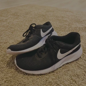 Svarta Nike sneakers strl 40 - Svarta Nike sneakers med vit sula och klassisk vit swoosh på sidan. Ovandelen är i mesh-liknande syntetmaterial som andas och har snörning framtill. Perfekta för dig som gillar sportig och clean stil. Storlek 40 (UK 6, US 7Y).